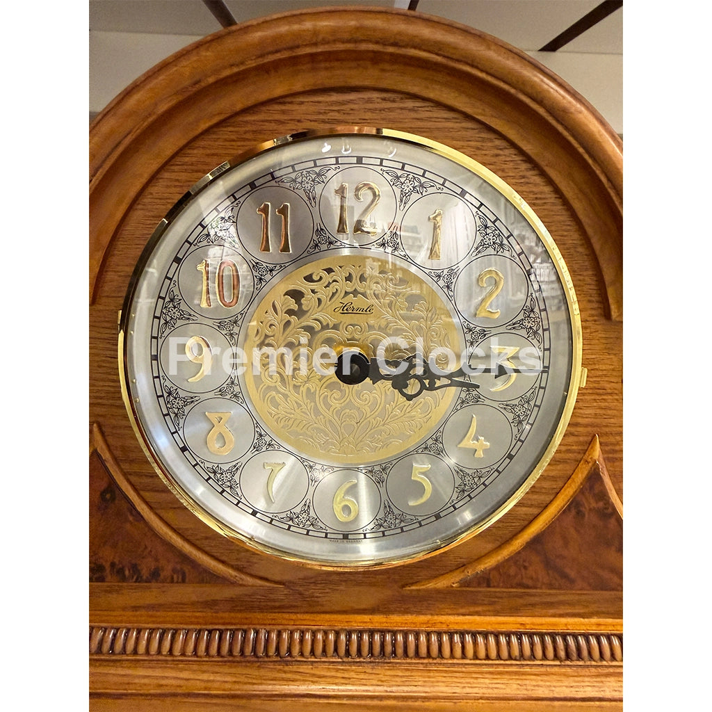 Hermle Amelia Mantel Clock in Light Oak real photo of a close-up of a wooden mantel clock face with a round silver and gold dial, ornate black hands, and a warm brown wood case. A semi-transparent 'Premier Clocks' watermark is visible across the center - Premier Clocks