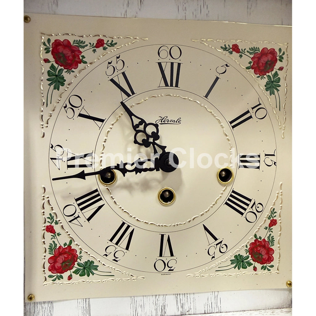 Hermle Austen Mechanical Mantel Clock in White real photo of an antique-style wall clock with a cream-colored face, black Roman numerals, ornate black hands, and red floral decorations in the corners - Premier Clocks