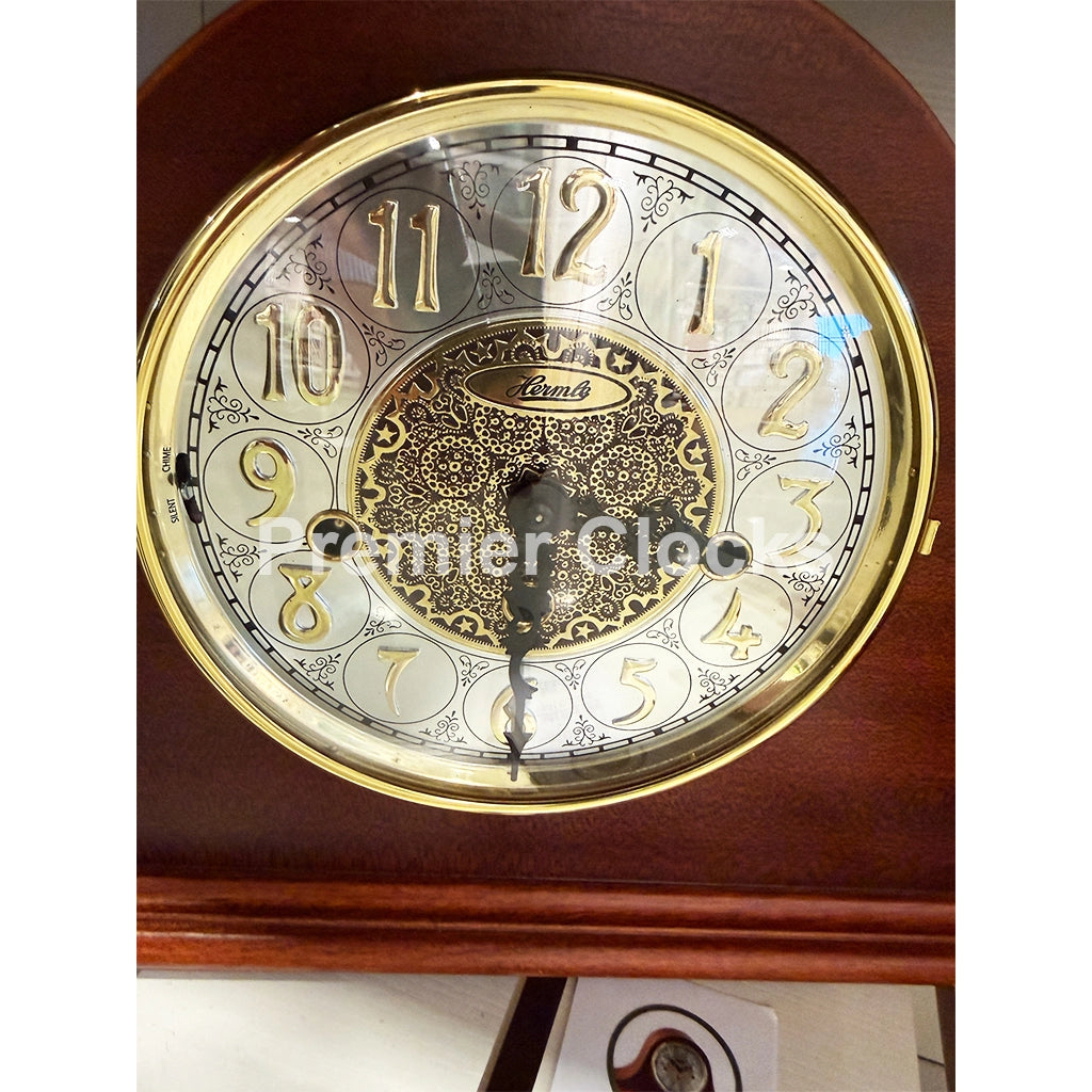Hermle Laurel Mantel Clock in Cherry is a close-up of a round antique-style wall clock face with a polished gold-tone bezel, ornate silver and gold numerals, and a central decorative medallion. The brand name 'Burlington' is visible on the center medallion - Premier Clocks