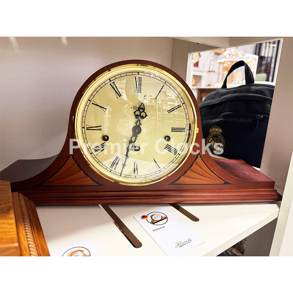 Hermle Atlanta photos and videos real photo of a wooden mantel clock with a cream-colored face, black Roman numerals, and gold-toned trim, displayed on a shelf with a branded card in front - Premier Clocks