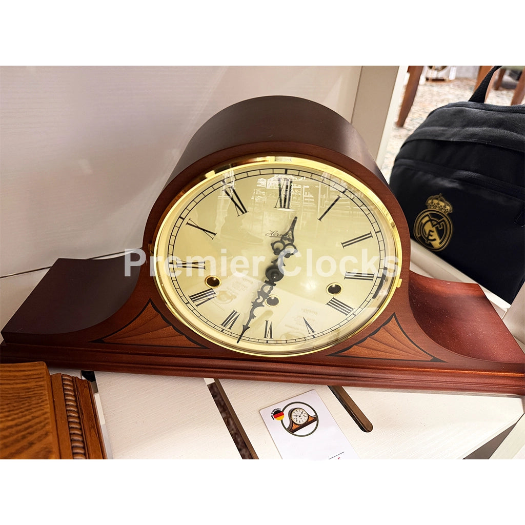 Hermle Remington Mantel Clock real photo of a wooden mantel clock with a cream-colored dial, gold-toned bezel, and dark brown wood casing, shown on a tabletop with a branded bag in the background - Premier Clocks
