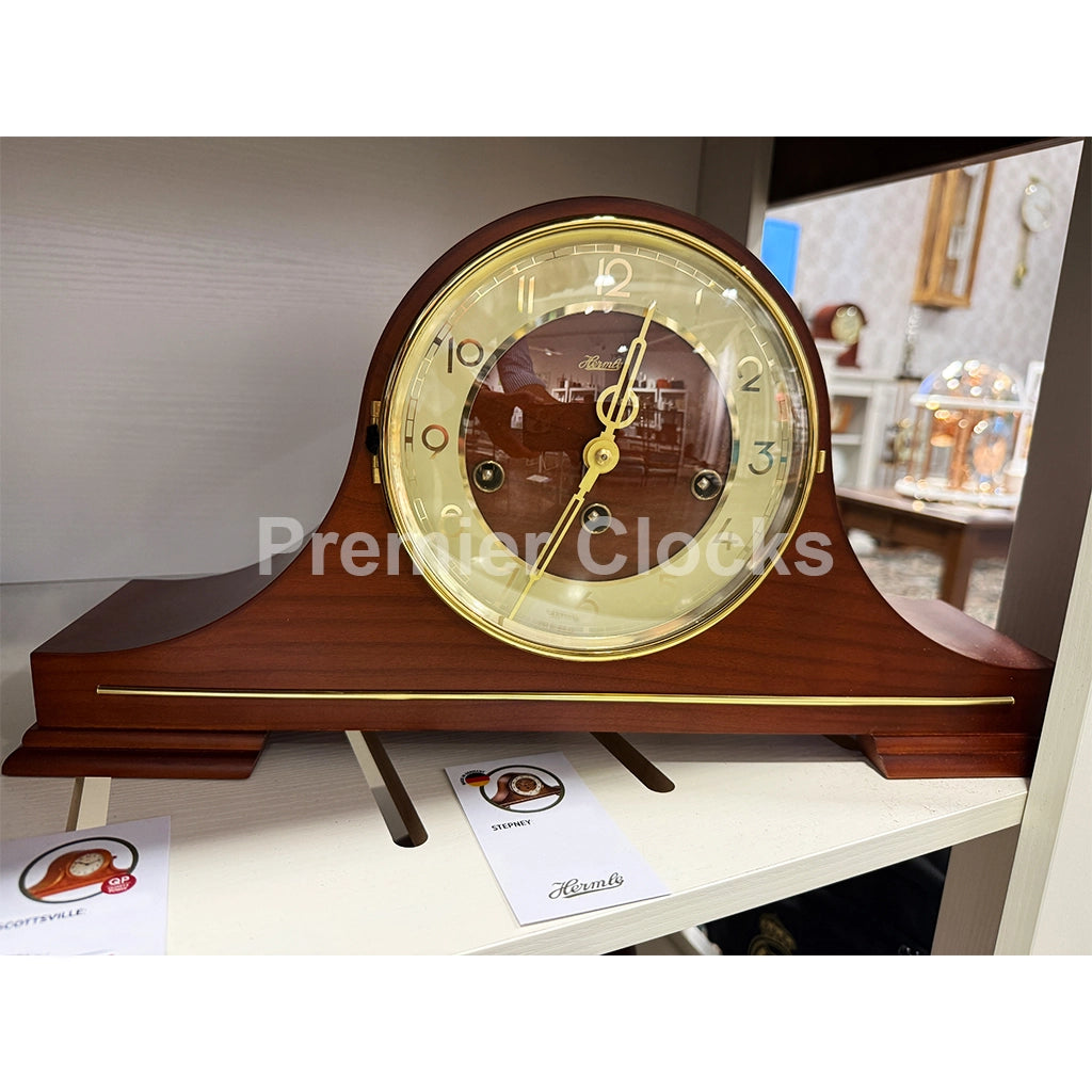 Hermle Stepney Mechanical Mantel Clock real photo of a wooden mantel clock with a warm brown wood finish, gold-tone hands and trim, and a cream-colored dial. The clock is displayed on a white shelf with two brand cards in front of it - Premier Clocks