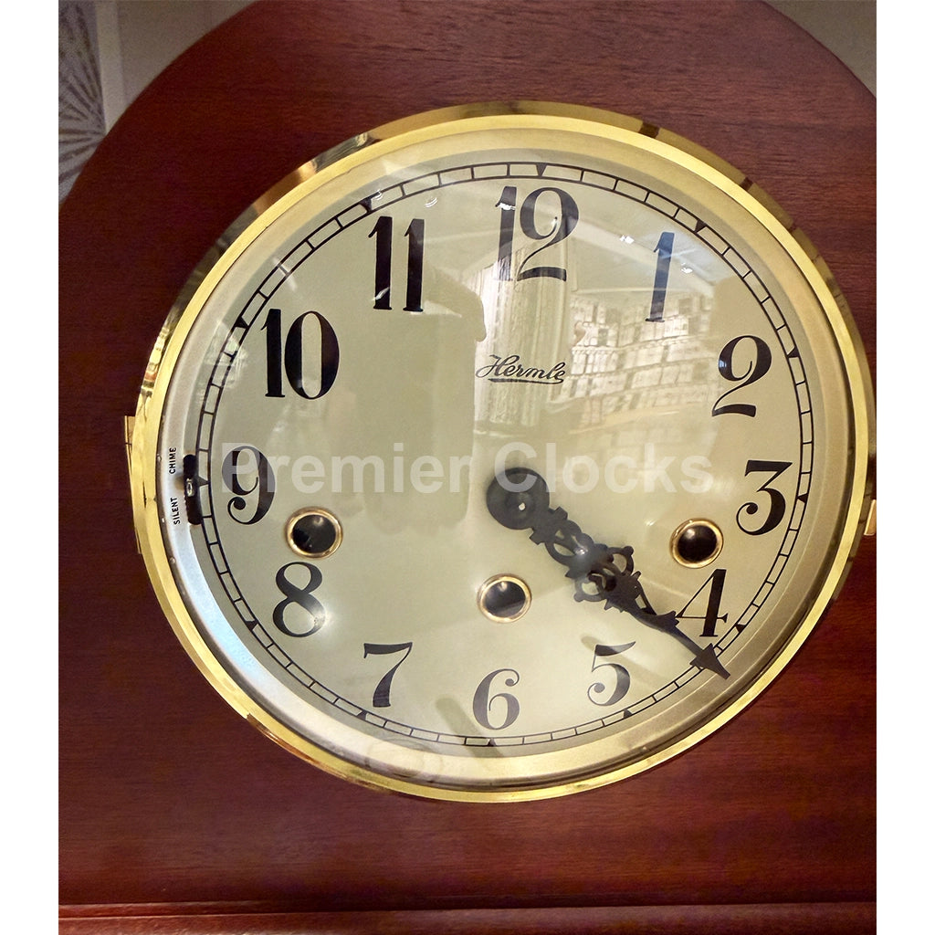Hermle Sweet Briar Mechanical Mantel Clock in Cherry is a close-up of a round clock face with a polished gold-tone bezel, black Arabic numerals, and black hands on a dark reddish-brown wooden mantel. The brand name 'Hermle' is visible on the clock face - Premier Clocks