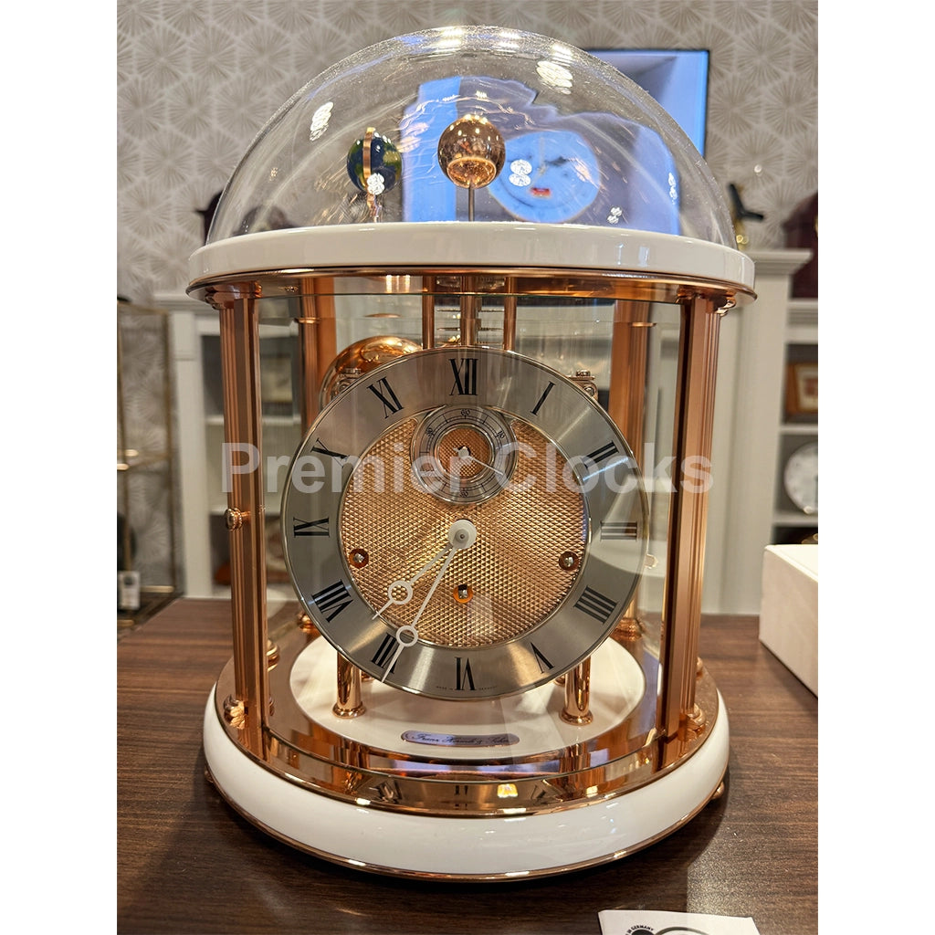 Hermle Tellurium Mantel Clock in White/Rose Gold real photo of a luxury-style table clock with a transparent glass dome, rose gold metal frame, white accents, and a round clock face with Roman numerals, displayed on a wooden table - Premier Clocks