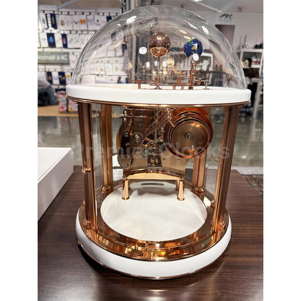 Hermle Tellurium Mantel Clock in White/Rose Gold real photo of a decorative globe clock with a clear acrylic dome, white circular base and top platform, and rose gold metal frame and internal clockwork mechanism, shown on a wooden table - Premier Clocks
