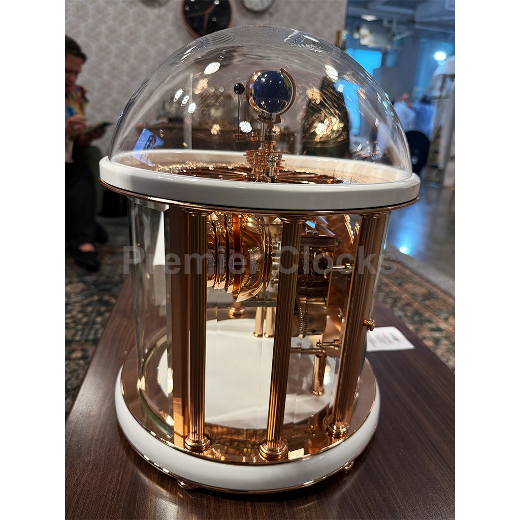 Hermle Tellurium Mantel Clock in White/Rose Gold real photo of a luxury mechanical clock under a clear glass dome display case, featuring a polished rose gold and white color scheme, set on a dark wooden table in an indoor showroom - Premier Clocks