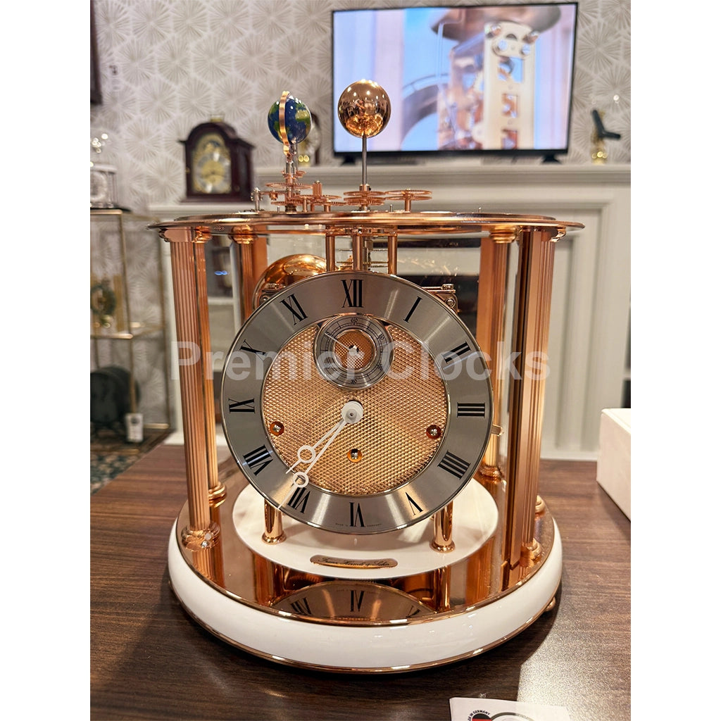Hermle Tellurium Mantel Clock in White/Rose Gold real photo of a luxury antique-style clock with a rose gold metal frame, a white marble base, and a silver-toned dial with Roman numerals, displayed on a wooden table in a home interior setting - Premier Clocks