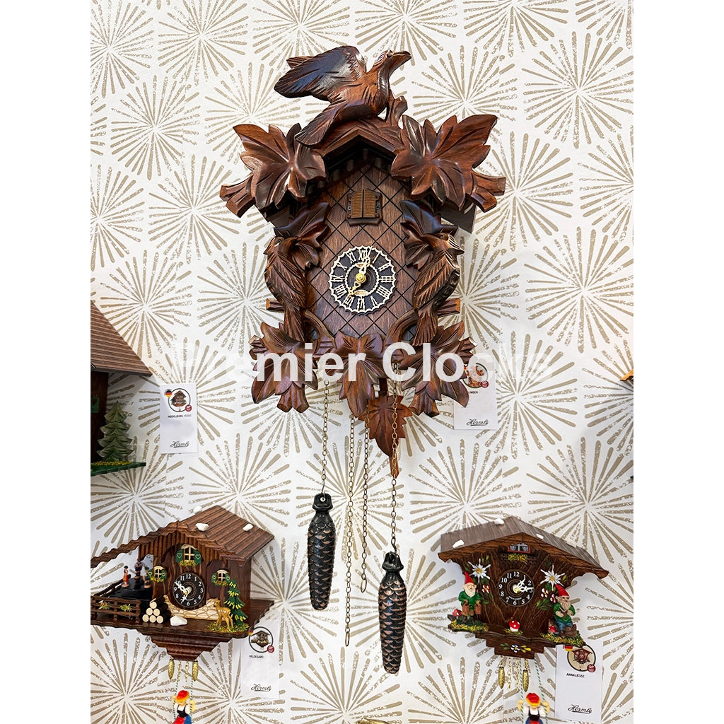 Hermle Villingen Cuckoo Clock is a large carved wooden cuckoo clock hangs on a patterned cream wallpaper background, with two smaller clocks below it. The main clock is dark brown wood with floral carvings, a clock face, bird figure on top, and hanging weights - Premier Clocks