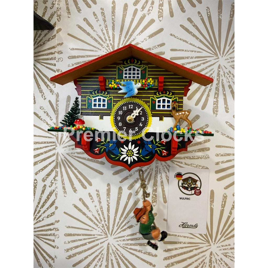Hermle Wulfric Cuckoo Clock real photo featuring a traditional black wood cuckoo clock shaped like a chalet house, featuring a colorful clock face, decorative roof, hanging gnome figure, and a brand card on a patterned background - Premier Clocks
